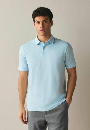 Mladý muž nosi svetlomodré polo tričko s krátkymi rukávmi a sivé nohavice, stojí s jednou rukou v vrecku pred jednoduchým pozadím.