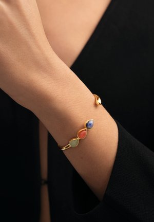 Muñeca con una pulsera de oro que tiene tres piedras en forma de lágrima en verde, rojo y azul, sobre una prenda de manga larga negra.