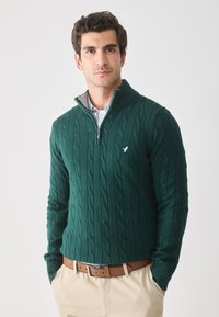 Man in een donkergroene kabelgebreide trui met kwartrits en een klein vogel logo, lichtblauw overhemd, beige broek en een bruine leren riem, met handen in de zakken.