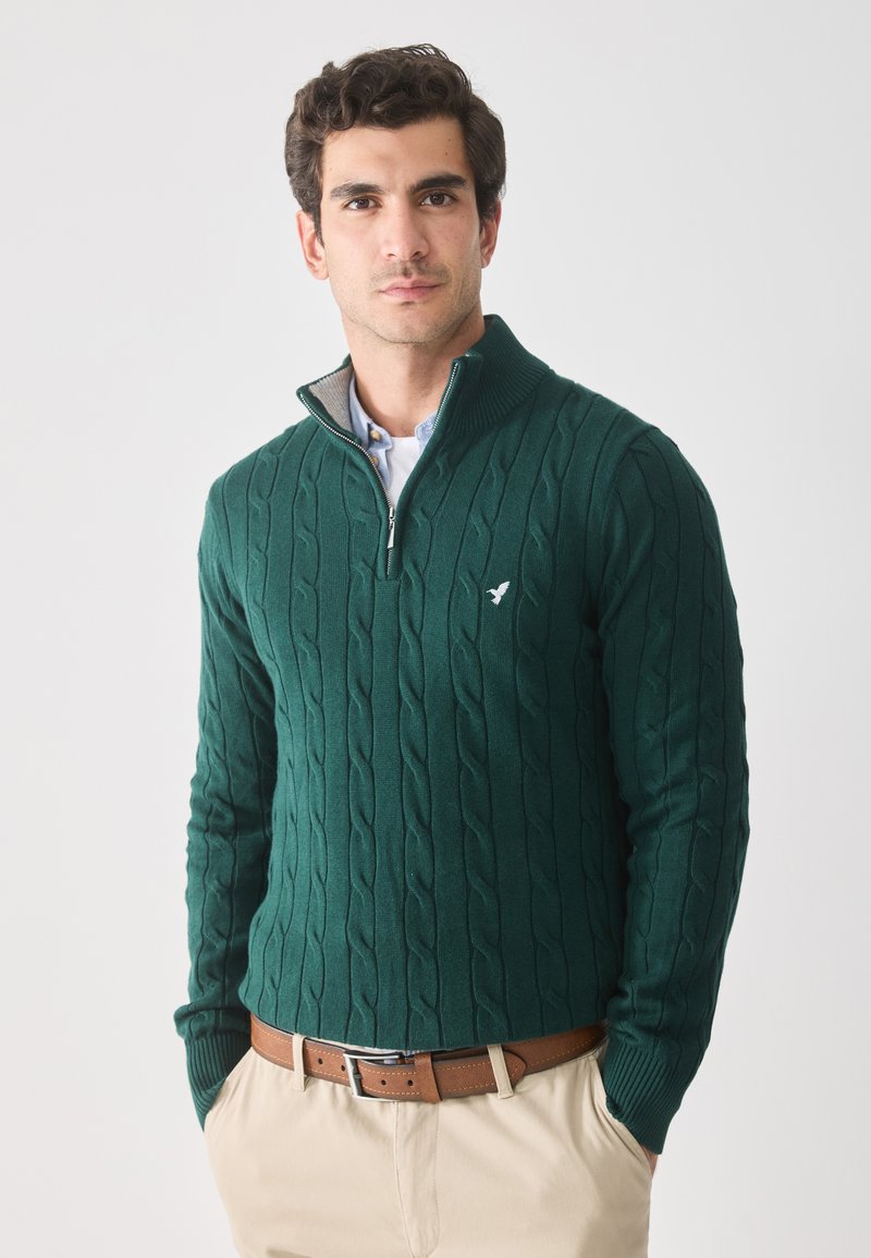 Man in een donkergroene kabelgebreide trui met kwartrits en een klein vogel logo, lichtblauw overhemd, beige broek en een bruine leren riem, met handen in de zakken.