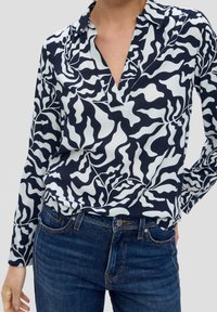 Person som bär en svart och vit blus med abstrakt mönster och lång ärm, instoppad i mörkblå denimjeans med knapp- och dragkedjeslutning.