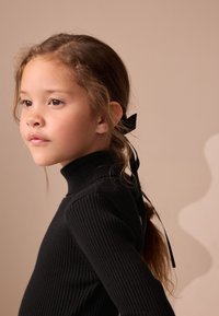 Jeune fille aux cheveux bruns attachés avec un ruban noir, portant un col roulé côtelé noir, regardant pensivement vers la gauche sur un fond beige.