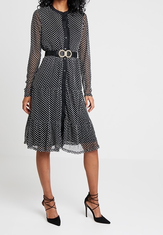 Femme portant une robe midi noire transparente à manches longues avec des pois blancs, une ceinture noire avec une boucle dorée, et des talons hauts noirs à lanières.