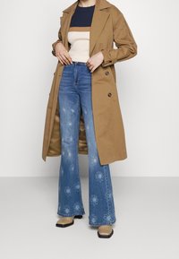 Kobieta ubrana w beżowy płaszcz trench, sweter w kolorowe bloki, niebieskie dżinsy z wysokim stanem w fasonie dzwonów z wzorem kwiatowym oraz beżowe botki z kwadratowym noskiem.
