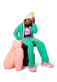 Groene trainingsbroek met witte en roze accenten, een marineblauwe T-shirt met grafisch ontwerp, een crèmekleurige gebreide muts met een gele pompon, en een roze gewatteerde jas.