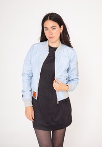 Ljusblå bomberjacka med en glänsande yta, grå ribbade manschetter och krage, dragkedja fram och en orange etikett på vänster sida.