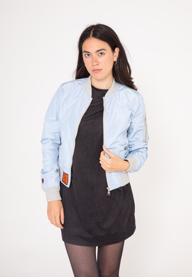 Ljusblå bomberjacka med en glänsande yta, grå ribbade manschetter och krage, dragkedja fram och en orange etikett på vänster sida.