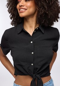 Camicia nera a maniche corte con bottoni e dettaglio annodato davanti. Presenta due tasche sul petto e un tessuto testurizzato.