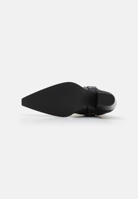 Zapato de tacón alto negro con punta afilada, suela texturizada y detalle de hebilla en el lateral, con un diseño elegante y un acabado brillante.