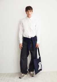 Weißes Hemd mit Kragen, dunkle Jeans, marineblauer Pulli um die Taille gebunden, graue und beige Turnschuhe und eine marineblaue Tragetasche mit weißem Logo.