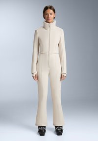 Beżowy kombinezon o pełnej długości z wysokim kołnierzem, zapinany na zamek z przodu, z długimi rękawami i teksturowanym wykończeniem, w połączeniu z czarnymi butami narciarskimi.