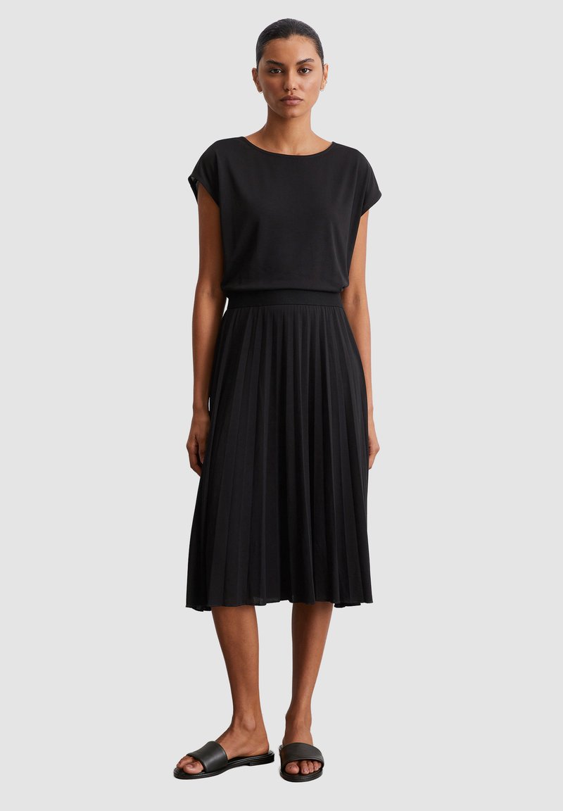 Zwarte plisséskirt gecombineerd met een zwart T-shirt met korte mouwen. Glad stof met een getailleerde taille en losse mouwen. Zwarte instapsandalen maken de look af.