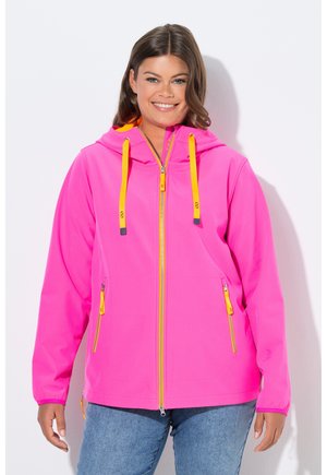 Lächelnde Frau, die einen leuchtend pinken Hoodie mit Reißverschluss, gelben Kordeln und Reißverschlussanhängern trägt, kombiniert mit blauen Jeans, vor weißem Hintergrund stehend.