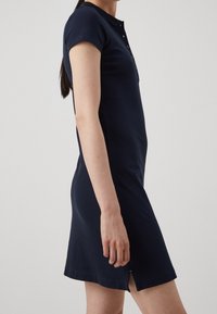 Vestido azul marino de manga corta con cuello tipo polo, tapeta de botones y slit laterales. La tela tiene una textura suave y una silueta ajustada.