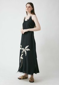 Schwarzes Maxikleid mit Spitzenbesatz am Ausschnitt, das auf der linken Seite ein beiges, gesticktes Palmenblatt-Muster und einen ungesäumten Saum aufweist.