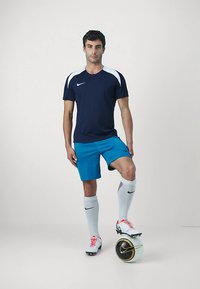 Mann i marineblå og hvite idrettsklær som står med en fot på en svart og hvit fotball foran en lys hvit bakgrunn.