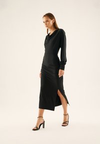 Black dress with long sleeves and cowl neckline, featuring ruching at waist, side slit, and a sleek, fitted silhouette. Paired with black heels.