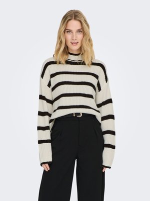 Ung kvinde med bølget blondt hår iført en cremefarvet sweater med sorte horisontale striber og sorte, højtaljede bukser med vide ben.