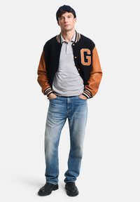 Veste varsity noire et brune avec un écusson, polo gris, jean bleu et chaussures noires ; fond blanc uni.