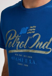 Modré bavlněné tričko s metalickým zlatým a modrým grafickým textem, včetně nápisu "Petrol Ind" a detailů o lokalitě, s kulatým výstřihem.