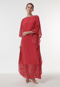 Rode maxidress met vleermuismouwen, voorzien van ingewikkelde lasergegoten kantdetails langs de randen. Vloeiende textuur, ontspannen pasvorm en een vloeiende silhouette.
