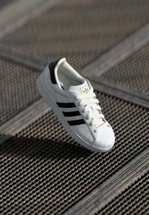 Biały sneaker z czarnymi paskami i noskiem typu shell unoszący się nad teksturowaną metalową powierzchnią w miękkim świetle.
