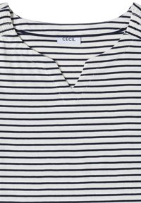 Chemise blanche avec des rayures horizontales noires, une petite encolure en V, un froncé élastique aux épaules et une étiquette de marque "CECIL" à l'intérieur du col.