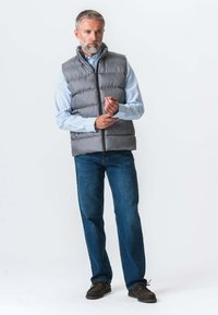 Grå polstret vest med høj krave, båret over en blå stribet skjorte. Modellen står i blå jeans og brune sko, mens han justerer skjortens manchetter.