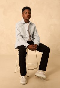 Light blue button-up shirt over a white t-shirt, black pants, and white sneakers. The subject sits on a metal stool against a beige background.