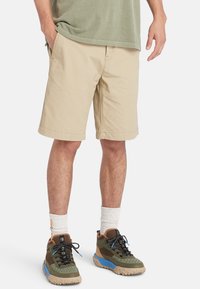 Pantalones cortos de algodón beige con bolsillos laterales, corte ajustado y dobladillo que cae justo por encima de la rodilla, combinados con zapatos de patrón oliva y marrón.