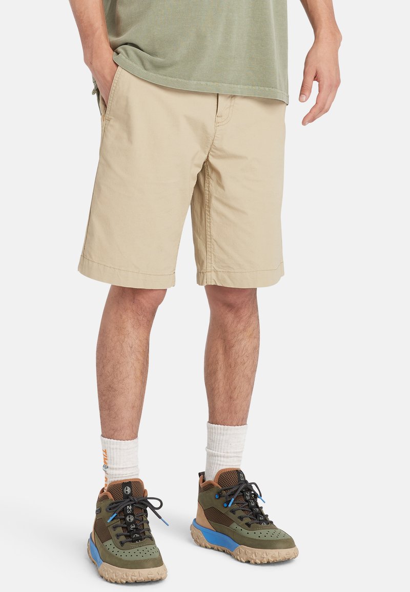 Beige shorts dari katun dengan saku samping, potongan yang disesuaikan, dan ujung yang jatuh tepat di atas lutut, dipadukan dengan sepatu berpola zaitun dan coklat.