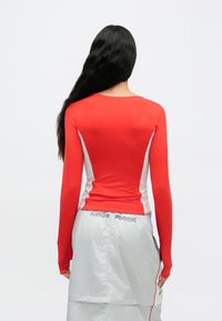 Czerwona bluzka z długim rękawem i białymi panelami po bokach, dopasowany fason, gładka tekstura, zestawiona z jasnoszarą spódnicą z logo "Nike Shox".