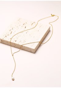 Goldbeschichtete Halskette mit einem gedrehten Kettendesign, ausgestattet mit einem einzelnen weißen Perlenanhänger. Ausgelegt auf einer strukturierten beigen Steinoberfläche.