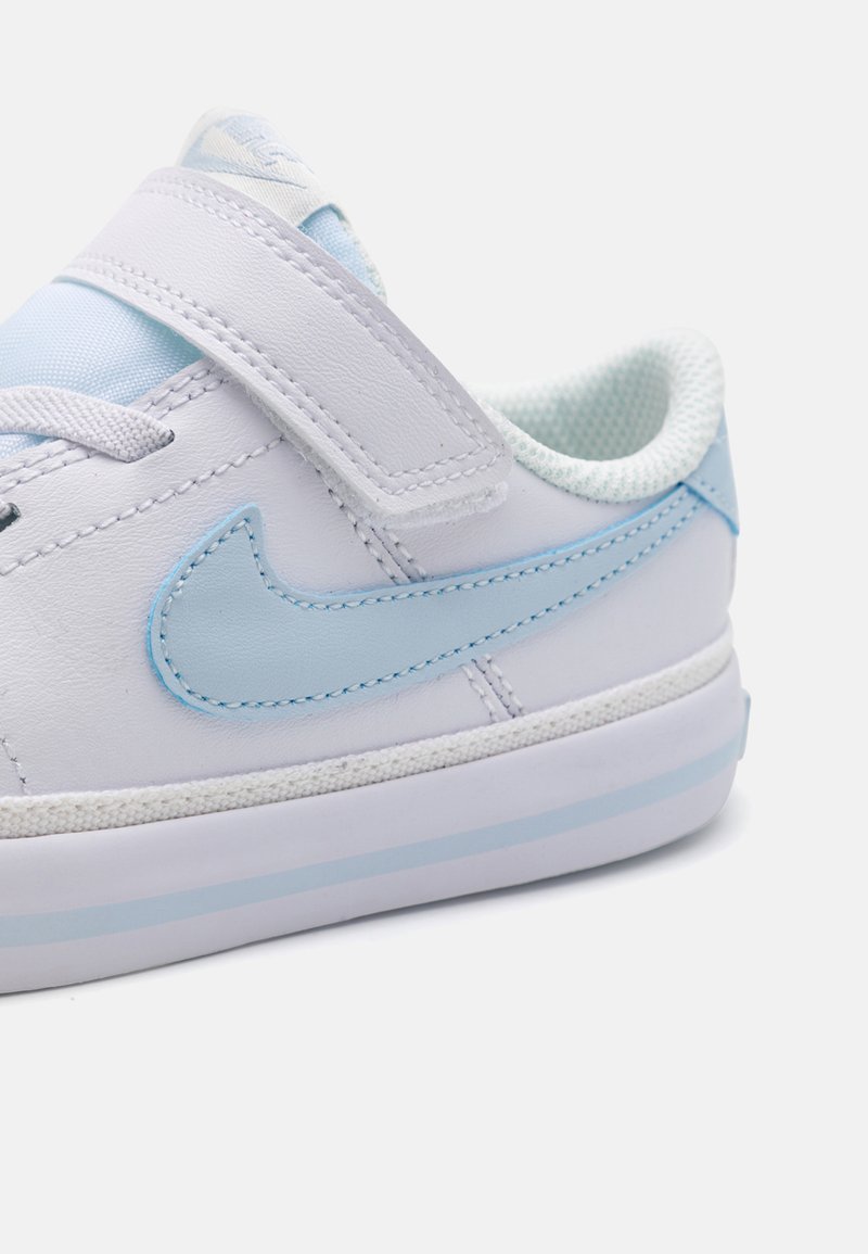 Zapatilla de piel blanca con acentos en azul claro y una correa de velcro. Presenta un interior texturizado y una suela de goma con rayas en azul claro.
