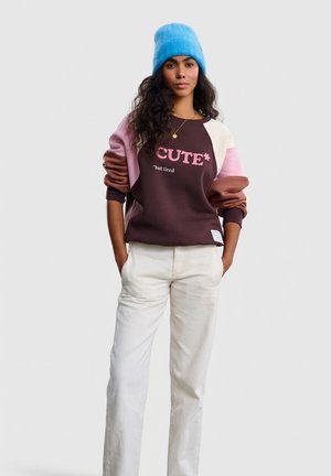 Bluza w kolorach brązowym, różowym i kremowym z napisem "CUTE*" oraz ściągaczami na rękawach. Połączona z białymi spodniami o prostych nogawkach. Modelka nosi niebieską czapkę beanie.