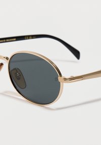 Occhiali da sole rotondi con montatura in metallo dorato e lenti grigie scure, con punte delle astine nere, su uno sfondo bianco.