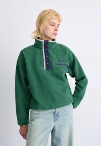 Pull en polaire vert avec un col haut, des boutons bleu marine et une poche poitrine zippée. Le tissu a une apparence texturée et une coupe décontractée.