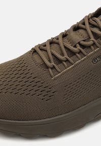 Zapatilla deportiva marrón con parte superior de malla, superficie texturizada, puntera reforzada y suela acolchada que presenta surcos prominentes para tracción.