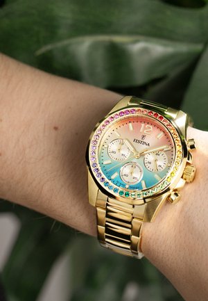Kultainen Festina-rannekello, jonka kehys on koristeltu sateenkaaren värisillä jalokivillä, tausta sinisestä vaaleanpunaiseen liukuvärjätty, kolme pieniä taulua ja kultainen metallilinkkiranneke ranteessa.