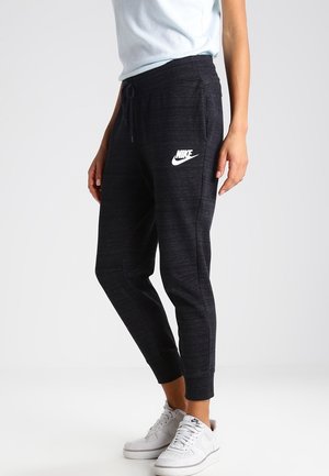 Osoba ubrana w czarne dresowe spodnie Nike i białe trampki Nike, stojąca na jednolitym jasnym tle.