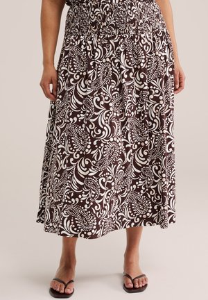 Mujer con falda midi marrón y blanca con estampado, cinturilla fruncida y sandalias negras de punta abierta, de pie frente a un fondo neutro.