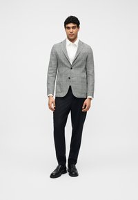 Άνδρας που στέκεται φορώντας γκρι καρό σακάκι, λευκό πουκάμισο, μαύρο παντελόνι και μαύρα παπούτσια, σε λευκό απλό φόντο.