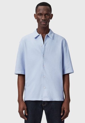 Mężczyzna na modelu ubrany w jasnoniebieską koszulę z krótkim rękawem na guziki i ciemne spodnie, stojący na jednolitym jasnym tle.