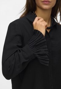 Black shirt featuring a placket front, collar, and gathered ribbed cuffs. Made of textured fabric with a soft finish.