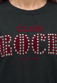 Μαύρο πουκάμισο με κόκκινο κείμενο "CLUB ROCK", διακοσμημένο με ασημένια τρουκς και μικρότερο κόκκινο κείμενο από κάτω που λέει "PLAY IT LOND"