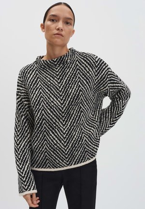 Vrouw in een zwart-wit zigzagpatroon trui met lange mouwen en zwarte broek, staand tegen een eenvoudige lichte achtergrond.