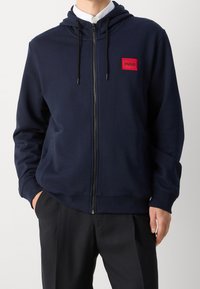 Hombre vestido con una sudadera con cremallera en color azul marino y un parche rojo con el logo "HUGO" en el pecho, y pantalones negros, de pie con las manos en los bolsillos.