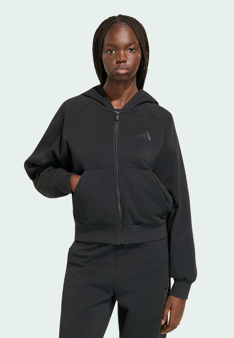 Jeune femme aux cheveux tressés portant un sweat à capuche noir zippé et un pantalon noir assorti, debout devant un fond clair.