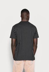 Černé tričko s krátkým rukávem s vertikálními bílými pruhy a kulatým výstřihem, vyrobené z lehkého bavlněného materiálu. Zobrazeno zezadu.