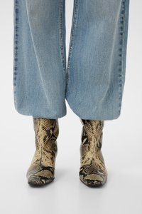 Jean large délavé clair porté sur des bottes mi-mollet à imprimé serpent avec des bouts pointus sur fond blanc.